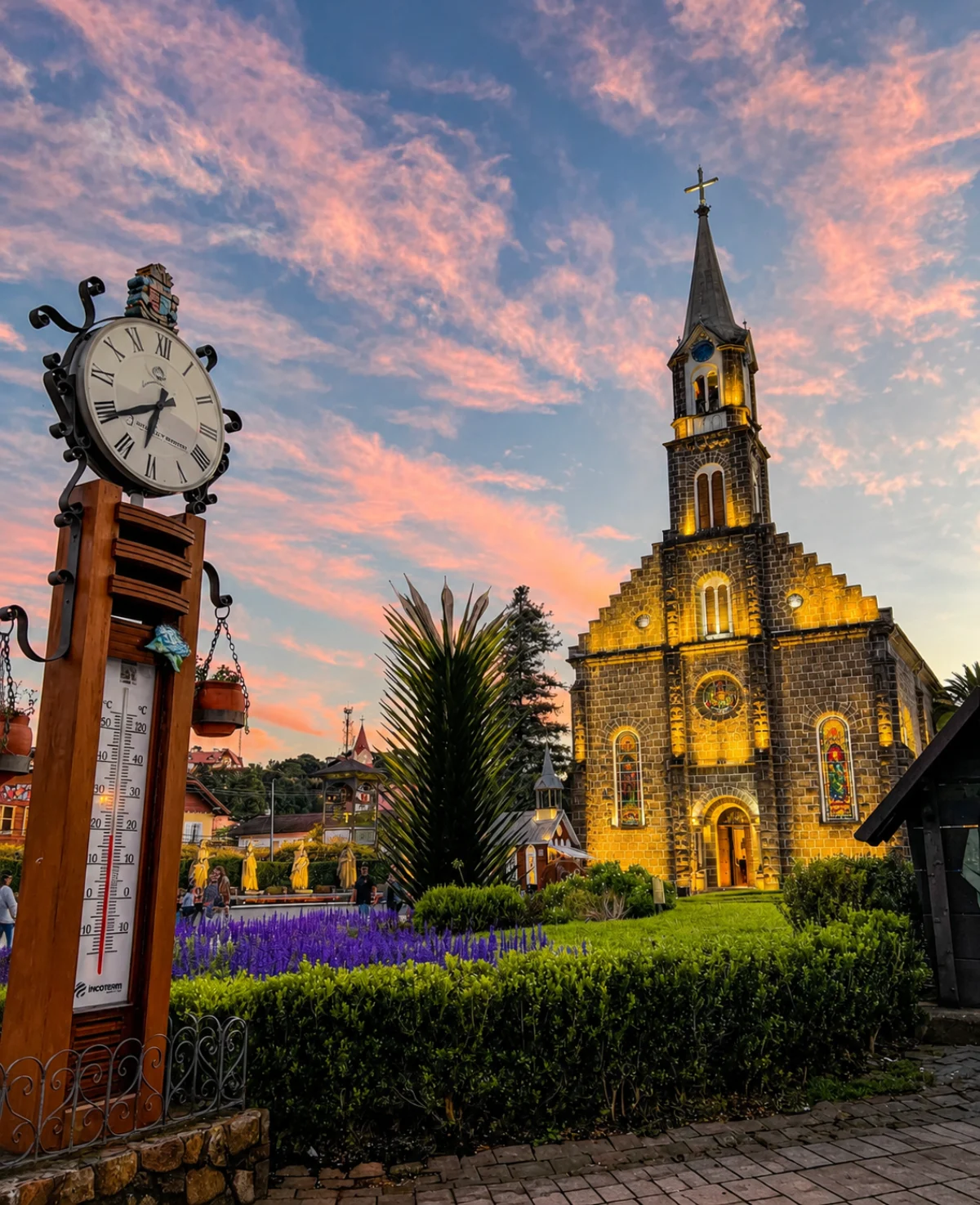 Torre da Igreja Matriz de Gramado com o relógio de rua e as lavandas em primeiro plano, sob um céu de pôr do sol rosa e azul